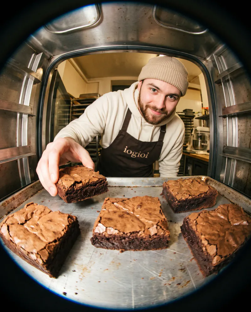 Fotografía con ojo de pez desde dentro de un horno: un pastelero con gorro y delantal de iogioi coge un brownie de la bandeja